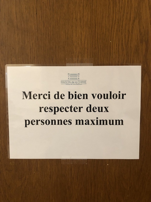 Une photo d'une feuille A4 collée sur une porte sur laquelle est écrit : 'Merci de bien vouloir respecter deux personnes maximum'.