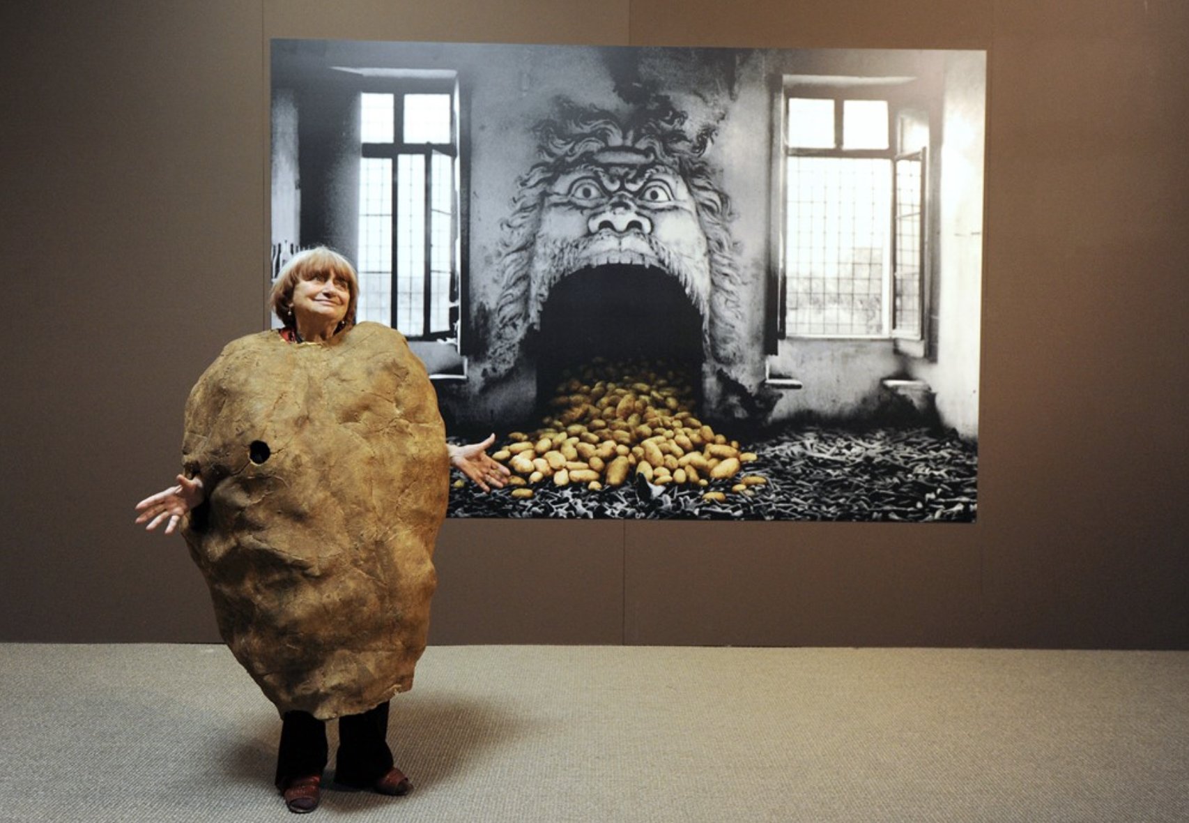 another photo of agnes varda dressed as a potato in front of a big photograph of a chimney looking like a face, potatoes in their mouth